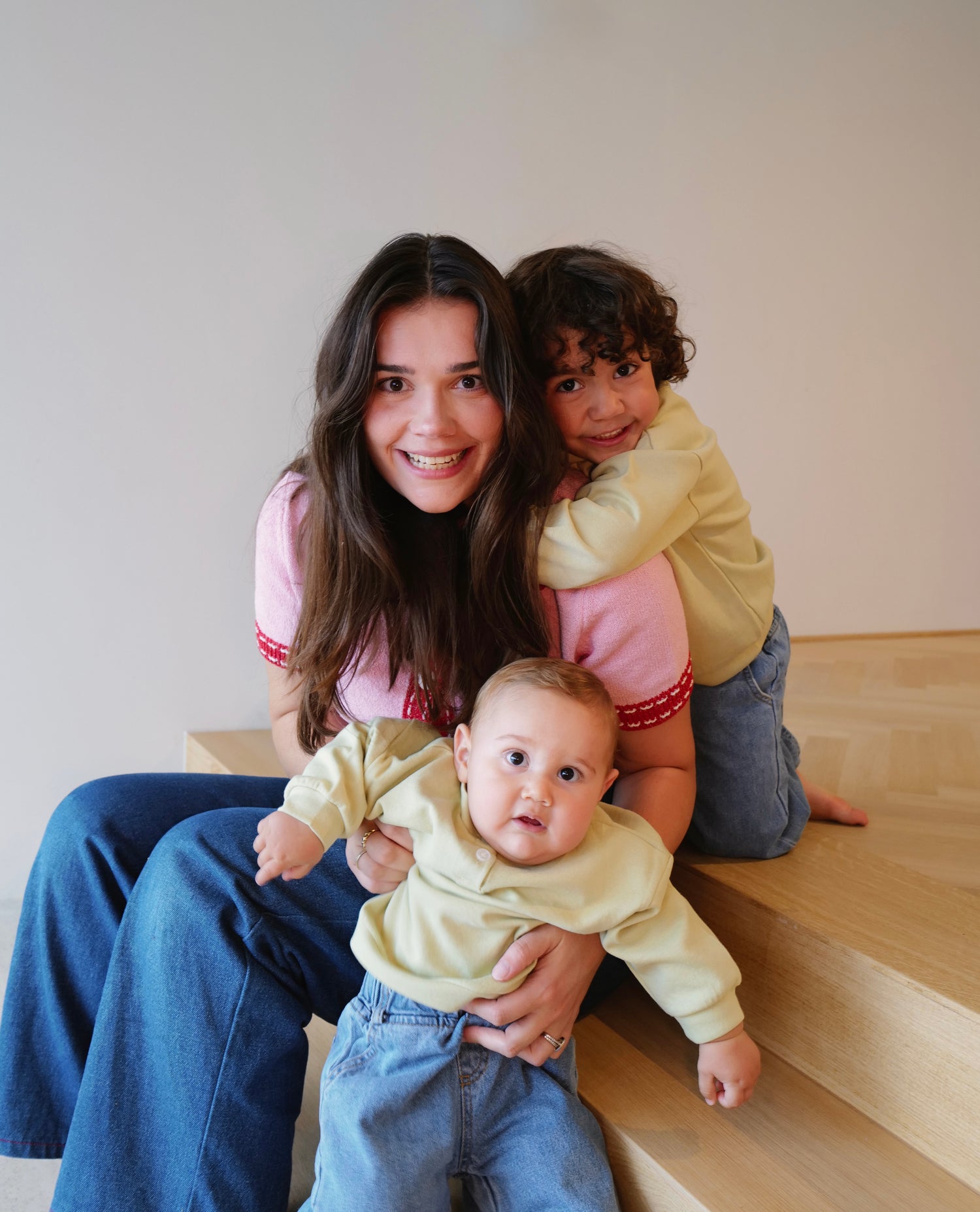 Woman sitting on a wooden floor with two children, one on her lap and another standing next to her.