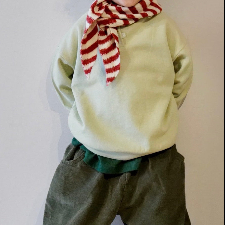 Child wearing a light green sweater and dark green pants with a red and white striped scarf, standing on a wooden floor.
