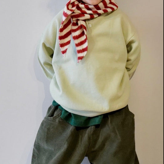 Child wearing a light green sweater and dark green pants with a red and white striped scarf, standing on a wooden floor.