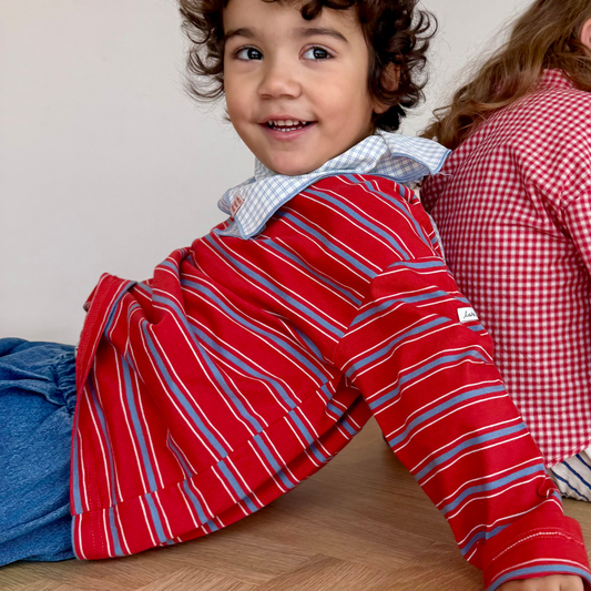 Child wearing a red striped shirt sitting on a wooden floor with a white wall in the background.
