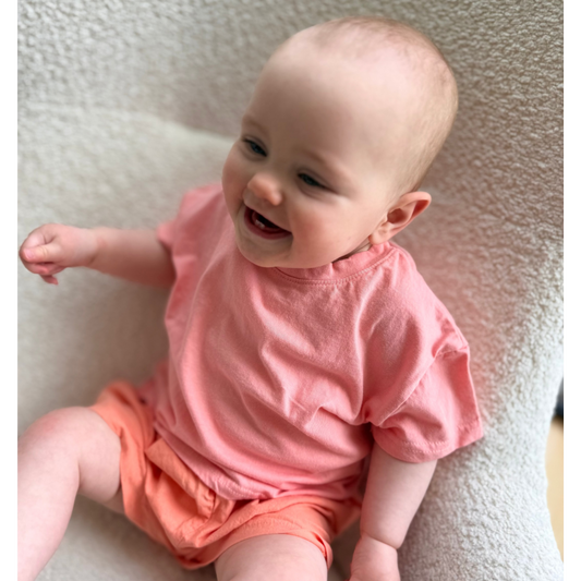 Baby wearing a pink shirt and orange shorts sitting on a textured surface
