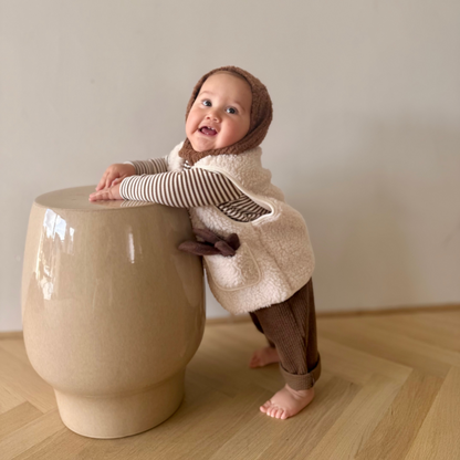 Child in a cozy outfit standing next to a round beige stool on a wooden floor.