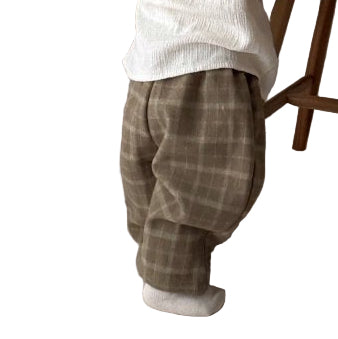 Child in a white sweater and plaid pants reaching up to a wooden chair with a pillow on it.